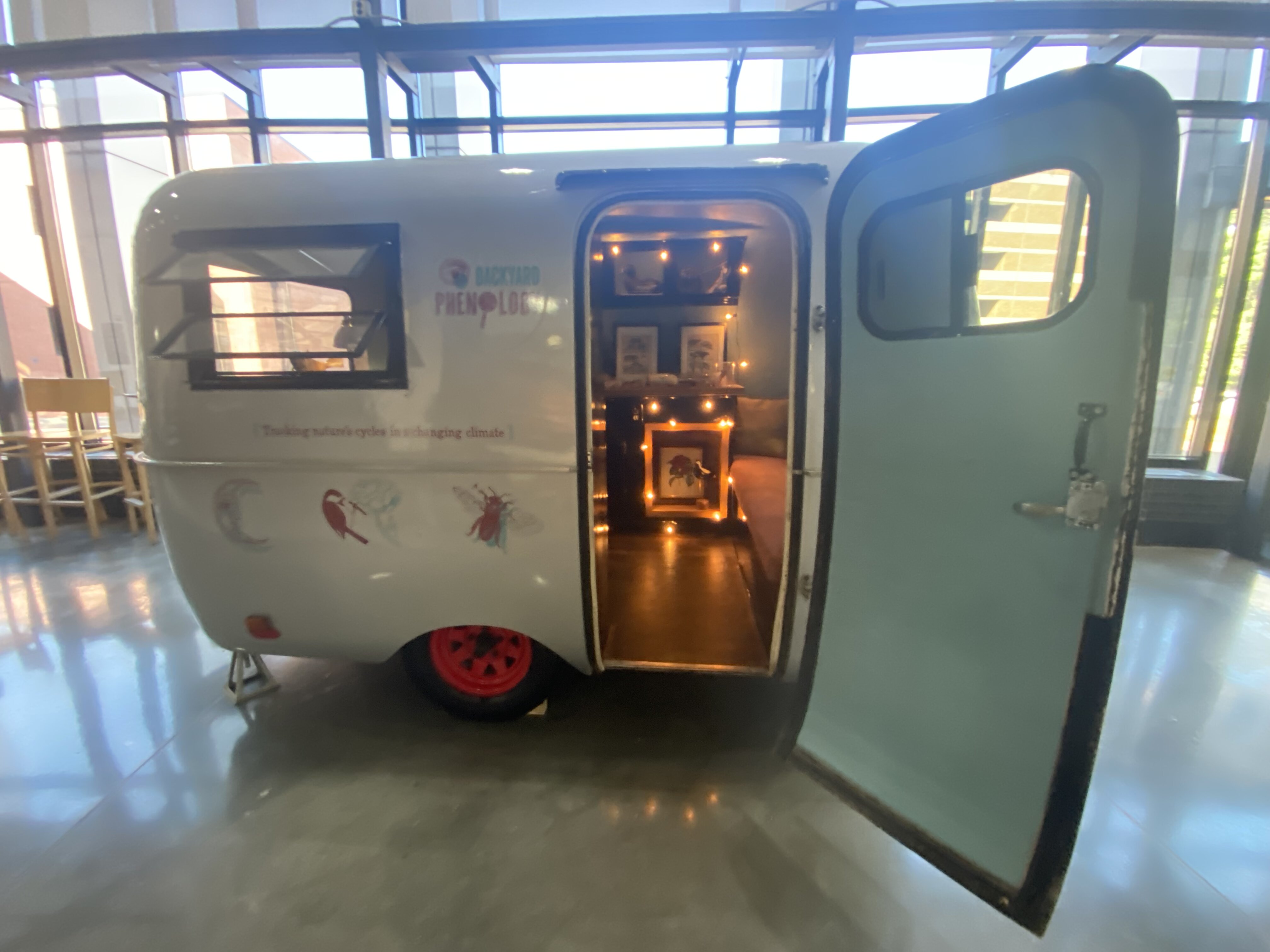 Silver camper in a gallery space, the door is open and interior lights welcome visitors