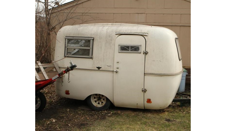 An castoff camper trailer with a rounded shape sits outside of a garage