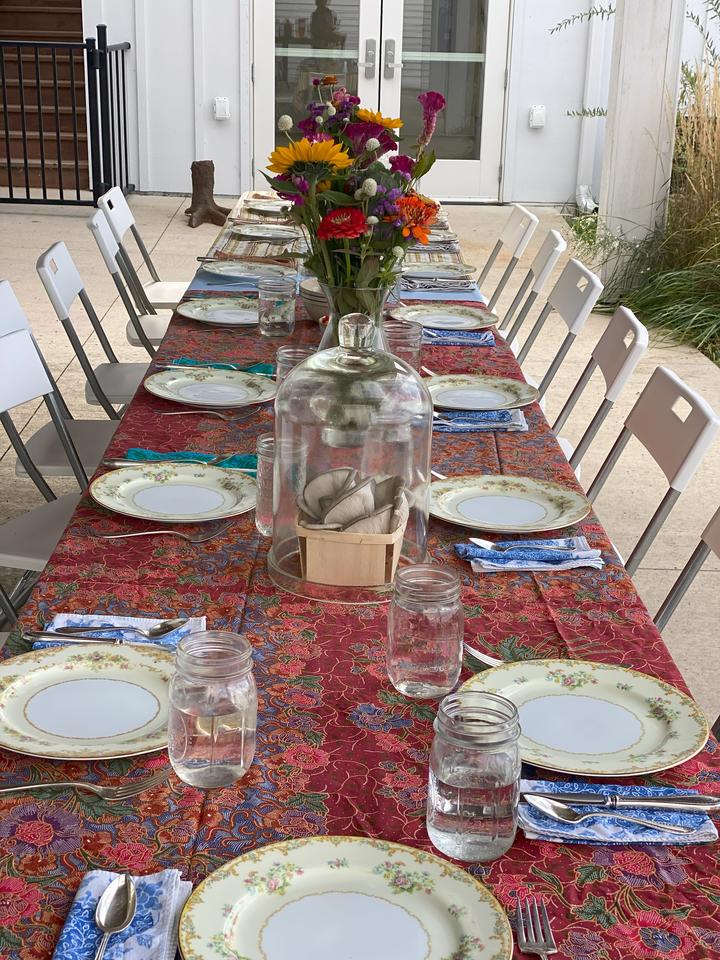 A long table with fifteen place settings. The colorful scene includes a red tablecloth, a large bouquet of colorful summer flowers, and living mushrooms under glass bell jars.