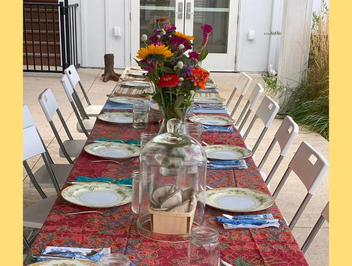 A long table set for a meal, red table cloth, white plates, blue napkins, and a vase of colorful flowers